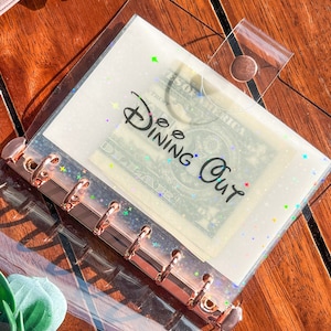 May include: Clear plastic binder with a rose gold ring mechanism. The binder has a clear pocket on the front with a dollar bill inside. The pocket has a glittery background and the text "Dee Dining Out" printed on it.