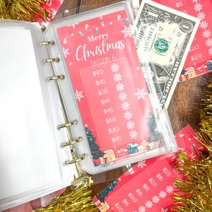 Pode incluir: Um fich&aacute;rio de pl&aacute;stico transparente com um inserto de tema natalino vermelho e branco. O inserto tem um t&iacute;tulo que diz "Feliz Natal Se&ccedil;&atilde;o 6" e lista v&aacute;rios valores em euros. Uma nota de um euro &eacute; vis&iacute;vel no fich&aacute;rio.