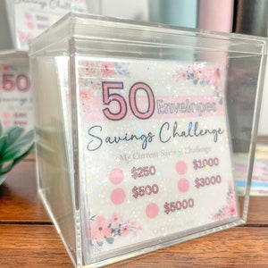 May include: A clear plastic box filled with 50 pink envelopes with a floral design. The envelopes are part of a savings challenge with amounts of $250, $500, $1000, $3000, and $5000.