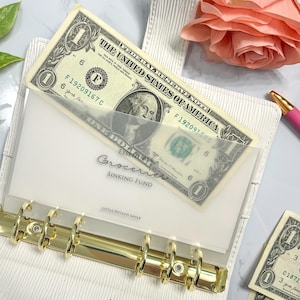 May include: A white planner with gold rings and a clear plastic pocket on the front. The pocket contains a one dollar bill with the text "The United States of America" and "One Dollar". The planner also has a blue ribbon tied around it. The text "Essentials Groceries Sinking Fund" is printed on the front of the planner.