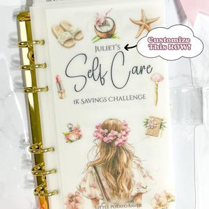 May include: A clear binder with gold rings and a decorative insert labelled "Self Care 1K Savings Challenge." The insert features illustrations of self-care items like slippers, a coconut, and a woman with flowers. The tab reads "SAVINGS HERE."