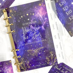 Peut inclure: Un classeur de défi d'épargne violet et doré avec le texte "Reach for the Stars" et "1K Savings Challenge". Le classeur a des anneaux dorés et des séparateurs mensuels. Des cartes supplémentaires avec des montants d'épargne sont visibles.