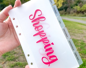 May include: A clear plastic binder with a pink "Shopping" title and dividers labeled "Shopping", "Savings", "Gas", "Groceries", "Household", and "Take Out".