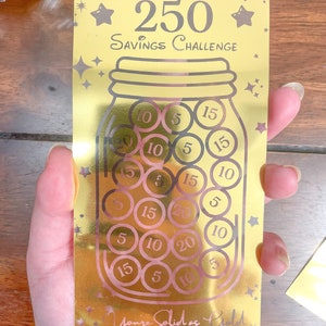 May include: A gold-colored card with a savings challenge theme. The card features a gold jar with pink circles inside, each with a number printed on it. The text on the card reads "Golden Jar 250 Savings Challenge" and "You're Solid as Gold Little Potato Soup".