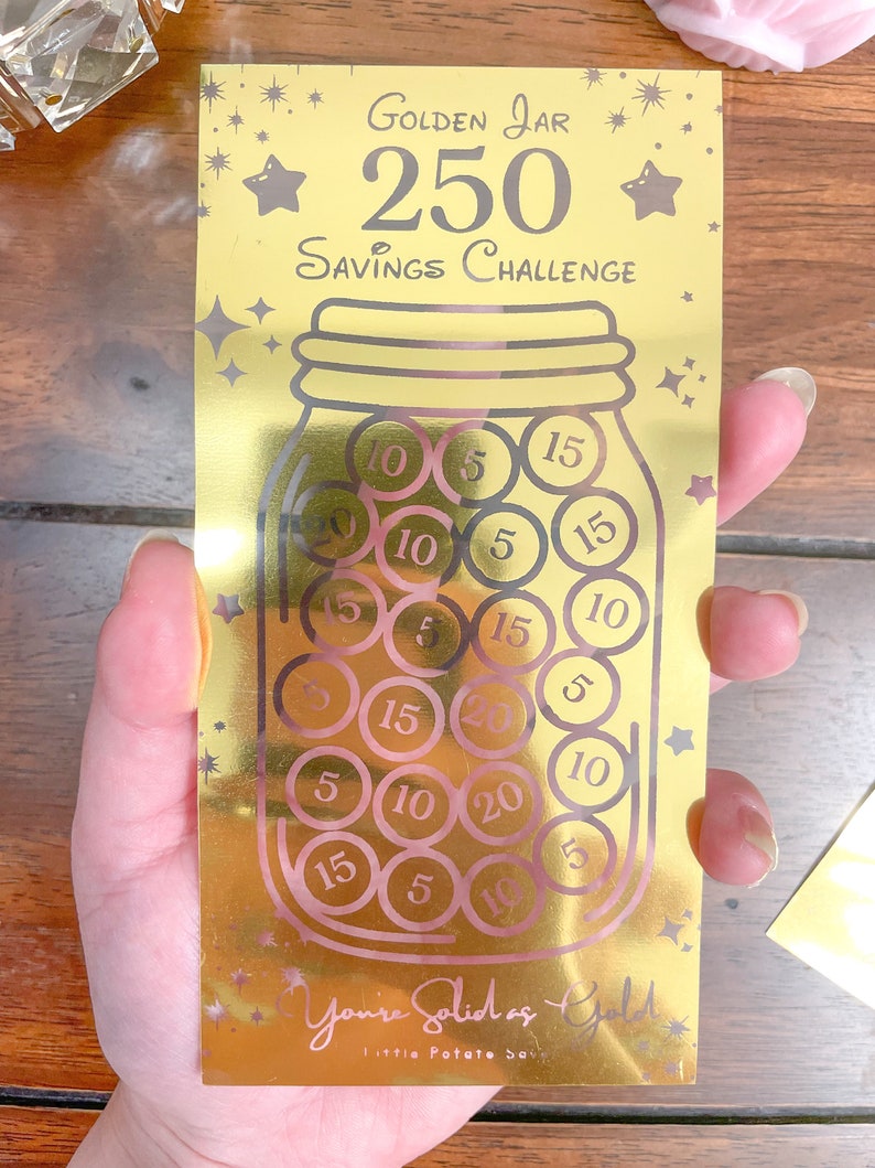 May include: A gold-colored card with a savings challenge theme. The card features a gold jar with pink circles inside, each with a number printed on it. The text on the card reads "Golden Jar 250 Savings Challenge" and "You're Solid as Gold Little Potato Soup".