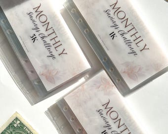 Op de afbeelding: Drie transparante plastic enveloppen met witte papieren inlegvelletjes. Elk inlegvelletje heeft een bloemmotief en de tekst "MONTHLY Savings Challenge" met een ander bedrag: 5K, 10K en 1K.