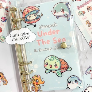 May include: A clear plastic binder with a blue and white cover featuring a seahorse, a turtle, a dolphin, and a clam. The cover has the text "Hannah Under The Sea" and "Ik Savings Challenge".