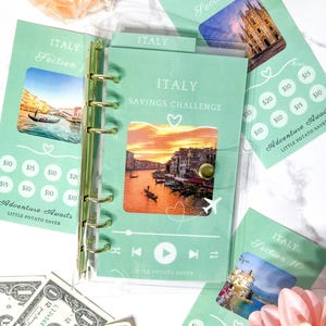 May include: A mint green six-ring binder labelled Italy Savings Challenge with a central photo of a canal at sunset, music player icons, and additional matching savings tracker inserts featuring Italian landmarks and currency amount circles.