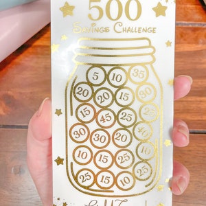 May include: A white card with gold lettering and stars. The card features a gold jar with gold coins inside. The coins have numbers on them, such as 5, 10, 15, 20, 25, 30, 35, and 40. The card reads "Golden Jar 500 Savings Challenge" and "Gold Coins! Little Potato Saver".