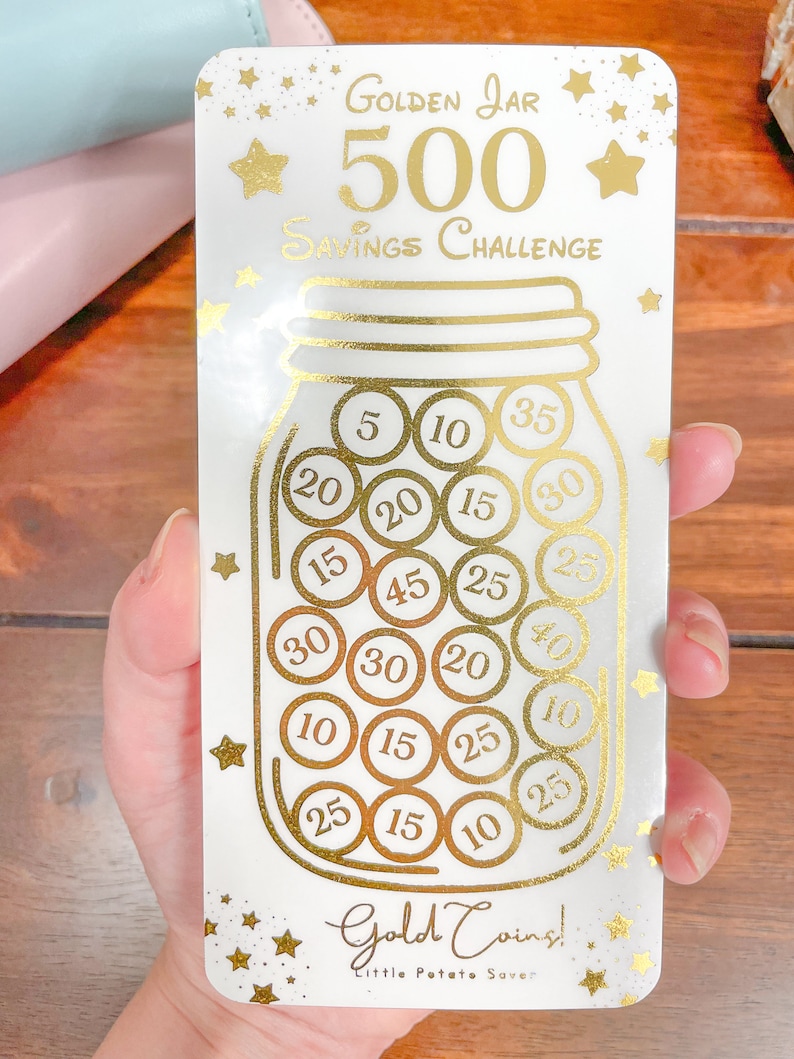 May include: A white card with gold lettering and stars. The card features a gold jar with gold coins inside. The coins have numbers on them, such as 5, 10, 15, 20, 25, 30, 35, and 40. The card reads "Golden Jar 500 Savings Challenge" and "Gold Coins! Little Potato Saver".