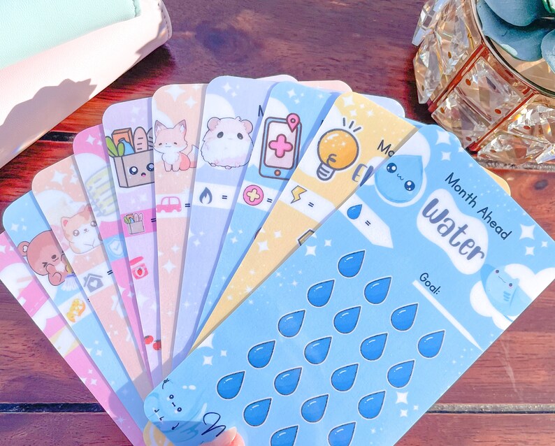 Pu&ograve; includere: Una collezione di colorate schede planner con simpatiche illustrazioni. Le schede presentano vari disegni, tra cui animali, cibo e icone. Una scheda ha il testo "Month Ahead Water" con grafiche a goccia d'acqua.