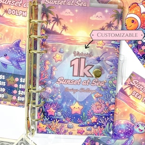 Peut inclure: Un classeur en plastique transparent avec des anneaux dorés contenant un insert de défi d'épargne Sunset at Sea. La couverture présente une scène océanique colorée avec des créatures marines, des étoiles et le texte Victoria's 1k Sunset at Sea Savings Challenge.