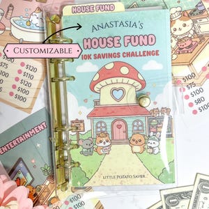 May include: A customizable binder with a gold spine, featuring a cover with a mushroom-shaped house illustration and the text "Anastasia's House Fund 10K Savings Challenge." Additional inserts include savings trackers and category labels like "Bathroom" and "Entertainment."