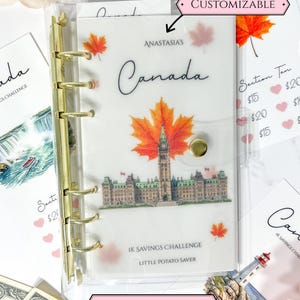 Peut inclure: Un classeur transparent avec une couverture en vélin mat doté d'un mécanisme à six anneaux dorés. La couverture affiche le texte "ANASTASIA'S Canada", "1K SAVINGS CHALLENGE" et "LITTLE POTATO SAVER" avec un motif du Parlement canadien et des feuilles d'érable.