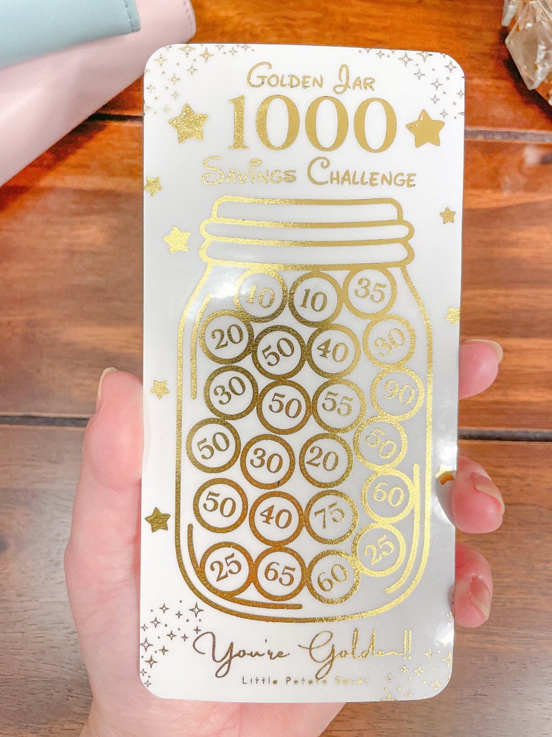 May include: A white card with gold lettering and a gold illustration of a jar filled with gold coins. The card says "Golden Jar 1000 Savings Challenge" and has numbers on the coins ranging from 10 to 90. The card also says "You're Golden!" and "Little Potato Seer".