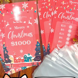 Pode incluir: Um rastreador de poupan&ccedil;a de Natal vermelho e branco com o texto "Feliz Natal 1000 &euro;" e uma ilustra&ccedil;&atilde;o festiva de uma cidade. O rastreador &eacute; acompanhado por envelopes de pl&aacute;stico transparente.