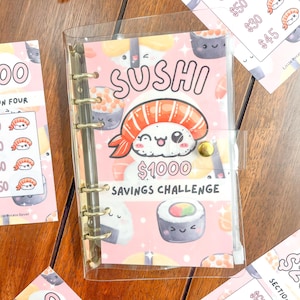 Puede incluir: Un archivador de plástico transparente con un interior rosa y blanco con un diseño de temática sushi. El archivador tiene anillas doradas y un título que dice "Sushi Savings Challenge". El interior presenta un rollo de sushi de dibujos animados y una pieza de sushi de dibujos animados con una cara sonriente. El archivador está abierto para revelar una página con un rollo de sushi de dibujos animados y el texto "1000 €".