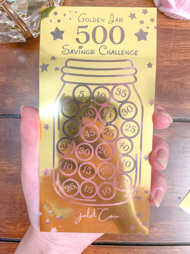 May include: A gold-colored card with a savings challenge theme. The card features a cartoon illustration of a glass jar filled with gold coins. The text "GOLDEN JAR 500 SAVINGS CHALLENGE" is printed on the card. The coins in the jar have different values printed on them, such as 5, 10, 15, 20, 25, 30, 35, and 40.