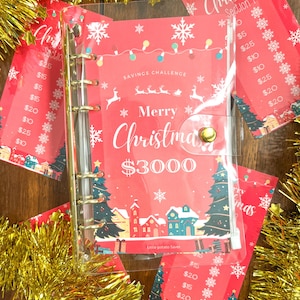 Pode incluir: Um fich&aacute;rio de pl&aacute;stico transparente com um inserto tem&aacute;tico de Natal vermelho e branco. O inserto tem um tema de desafio de poupan&ccedil;a com o texto "Feliz Natal 3000 &euro;" e uma imagem de uma pequena cidade com &aacute;rvores de Natal. O fich&aacute;rio tem argolas douradas e um bot&atilde;o dourado.