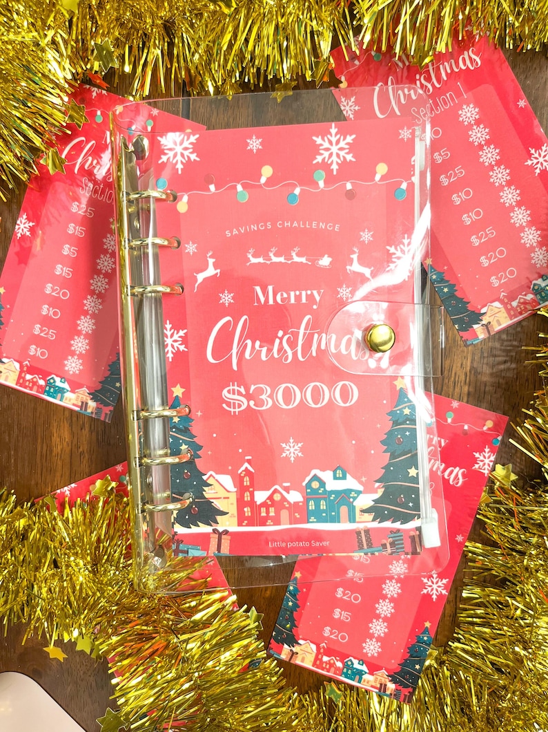 Pode incluir: Um fich&aacute;rio de pl&aacute;stico transparente com um inserto tem&aacute;tico de Natal vermelho e branco. O inserto tem um tema de desafio de poupan&ccedil;a com o texto "Feliz Natal 3000 &euro;" e uma imagem de uma pequena cidade com &aacute;rvores de Natal. O fich&aacute;rio tem argolas douradas e um bot&atilde;o dourado.