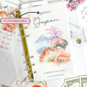 May include: A Japan-themed savings challenge binder with a matte vellum cover. The design features a geisha, Mount Fuji, and cherry blossoms. The binder includes gold rings and inserts for tracking savings.