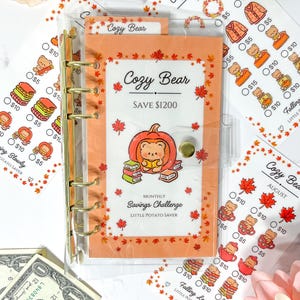 Peut inclure: Un classeur A6 transparent avec des anneaux dorés contenant une fiche de défi d'épargne mensuelle Cozy Bear. La carte orange présente un ours dans une citrouille, des livres et des feuilles d'érable avec le texte Save $1200. Des fiches montrent des ours.