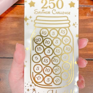 May include: A white card with gold lettering and a gold jar illustration. The card says "Golden Jar 250 Savings Challenge". The jar is filled with gold coins with numbers on them, such as 5, 10, 15, and 20. The card also says "You're Solid as Gold" and "Little Payam Studio".