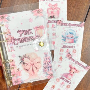 May include: A clear binder with gold rings and a snap closure, featuring pink Christmas-themed inserts. The inserts include illustrations of a gift, snow globe, and a deer, with text reading "Pink Christmas" and "5K Savings Challenge."