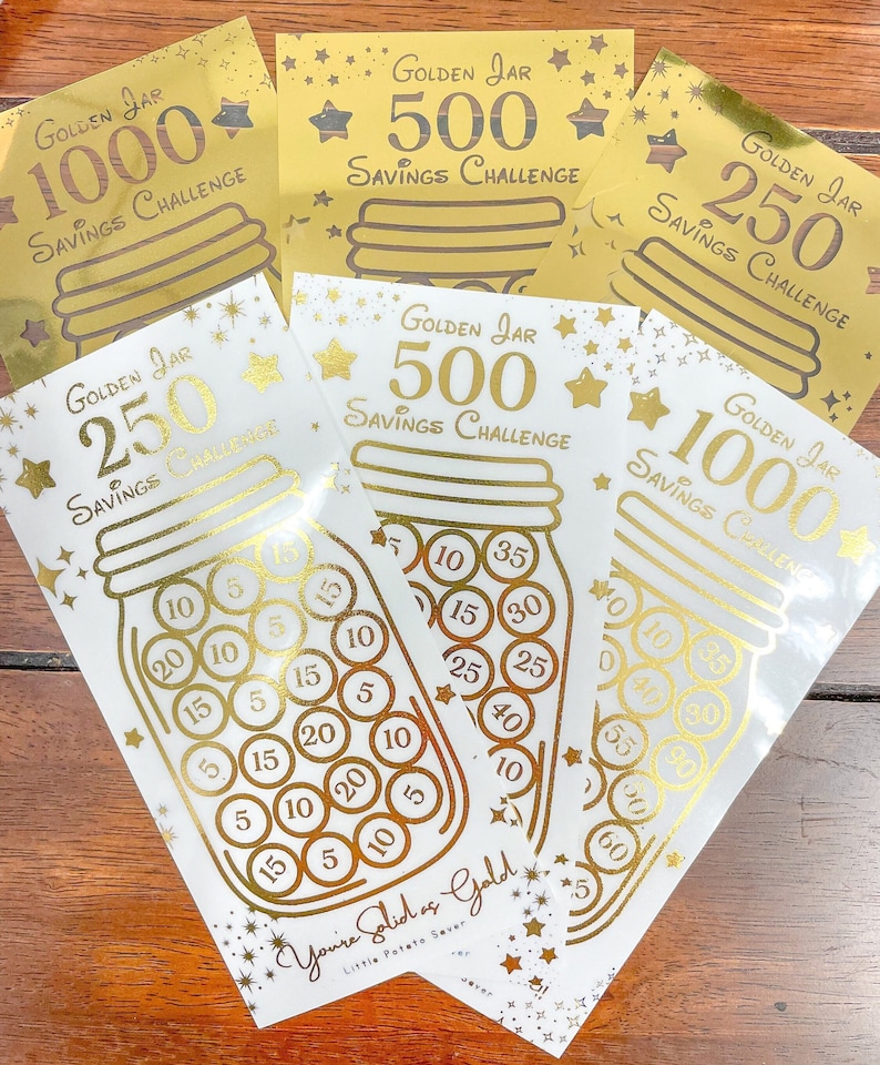 May include: Five gold foil stickers with a jar design and the text "Golden Jar Savings Challenge" with different amounts: 1000, 500, 250, 500, and 1000. The stickers have a gold glitter background and stars.