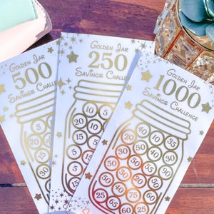 May include: Three gold foil printed cards with a savings challenge theme. Each card features a mason jar with coins inside and a gold star. The cards are labeled "Golden Jar 500 Savings Challenge", "Golden Jar 250 Savings Challenge", and "Golden Jar 1000 Savings Challenge".