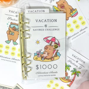 Peut inclure: Un classeur en plastique transparent avec des anneaux dorés contenant un encart Vacation Savings Challenge. La couverture présente un capybara de dessin animé sous un parasol avec le texte $1000 Adventure Awaits Little Potato Saver. Des encarts supplémentaires avec des illustrations de capybara sont visibles.
