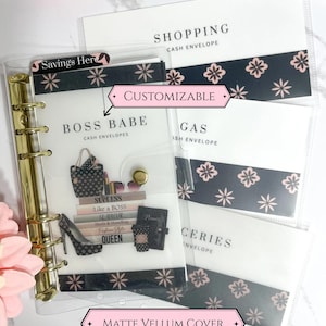 May include: A clear binder with gold hardware and several cash envelopes. The envelopes are labeled "Shopping", "Gas", and "Groceries". The binder has a "Boss Babe" cover with illustrations and the text "Savings Here", "Customizable", and "Matte Vellum Cover".