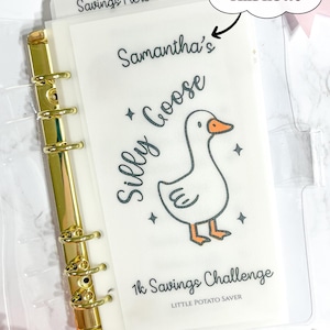 Peut inclure: Un classeur transparent avec une tranche dorée et un insert de défi d'épargne. L'insert présente une oie de dessin animé avec le texte "Samantha's Silly Goose" et "1k Savings Challenge". L'onglet indique "Savings Here".