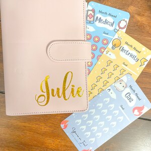 Pu&ograve; includere: Un'agenda rosa chiaro con il nome "Julie" in oro. L'agenda &egrave; accanto a tre schede di budget con le etichette "Medical", "Electricity" e "Gas". La scheda "Gas" ha il testo "You're on Fire!" e una piccola grafica a fiamma.