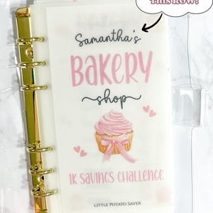 Peut inclure: Un classeur transparent avec une reliure dorée et un insert rose et blanc. L'insert présente le texte "Samantha's Bakery Shop" et une illustration de cupcake. Le texte "1K Savings Challenge" est également visible. L'onglet indique "Savings Here".