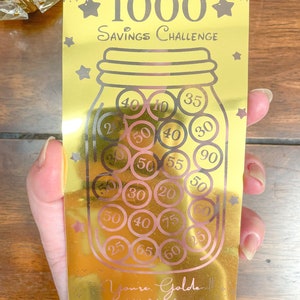 May include: Gold savings challenge jar with a gold background and a gold lid. The jar is filled with pink and black coins with numbers on them. The text on the jar reads "Golden Jar 1000 Savings Challenge".