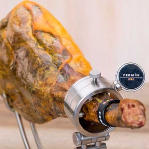 May include: A whole Iberian ham, held in a stainless steel ham holder. The ham is a rich brown color with visible marbling. The holder has a circular clamp and a label that reads "FERMÍN".