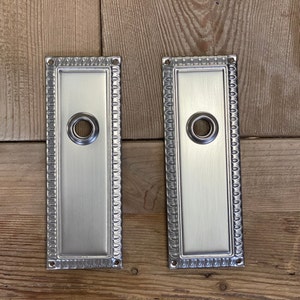 May include: Two rectangular, brushed silver door plates with a decorative beaded border and a circular hole in the center. The plates are set against a wooden background.