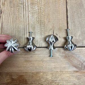 May include: Four silver cabinet knobs with a decorative, textured design. The knobs are arranged in a row on a wooden surface.