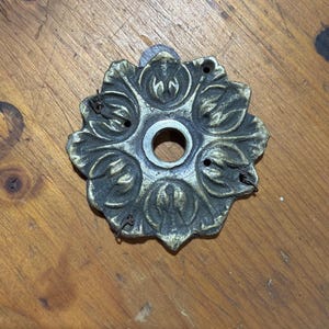 May include: A circular metal escutcheon plate featuring a raised floral petal design with a dark antique finish. The piece has a central circular opening and three small mounting holes around the perimeter, with thin wire remnants attached.