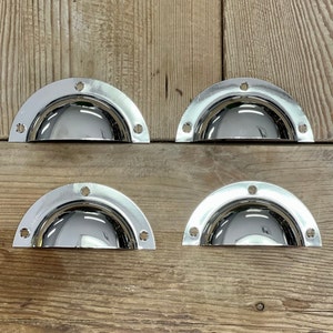 May include: Four chrome cup-shaped cabinet pulls with a domed design. The pulls are arranged in two rows of two on a wooden surface.