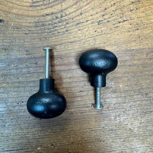 May include: Two black, cast iron cabinet knobs with a round, bulbous design. Each knob has a threaded post for mounting. The knobs are displayed on a wooden surface.