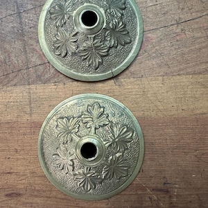 Puede incluir: Dos objetos circulares antiguos de latón con un agujero central y un diseño de hojas. Los objetos están sobre una superficie de madera. El latón tiene un aspecto ligeramente envejecido.