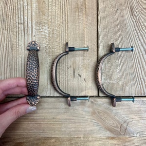 May include: Three copper-colored cabinet pulls and a matching handle rest on a wooden surface. The pulls are curved with a hammered texture, and the handle is oval with a similar hammered design. Each pull has a screw for installation.