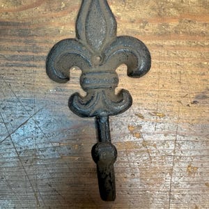 May include: A black fleur-de-lis wall hook made of cast iron. The hook has a decorative fleur-de-lis design at the top with a curved hook at the bottom. The hook is mounted on a wooden surface.