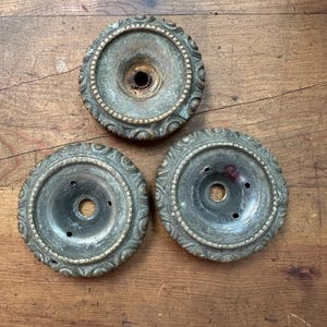 Puede incluir: Tres elementos decorativos circulares de metal antiguo con aspecto envejecido. Cada pieza presenta un orificio central y un borde ornamentado en relieve con un patrón detallado. El metal tiene un tono bronce verdoso deslustrado.