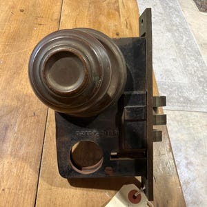 May include: A dark brown metal mortise lock set featuring a round knob and a rectangular faceplate with three protruding bolts. The metal casing displays the stamped text 551-2 3/4-112-L and includes a circular opening for a spindle mechanism.