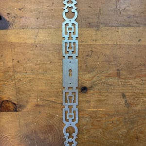 May include: A silver-colored, decorative metal door plate with an ornate, symmetrical design. The plate features geometric shapes and a keyhole, with decorative flourishes at the top and bottom. The plate is approximately 12 inches long.