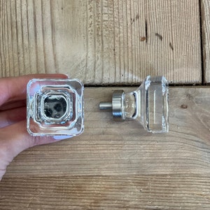 Może przedstawiać: Dwa kwadratowe, przezroczyste gałki meblowe ze srebrną podstawą. Gałki mają fazowane krawędzie.