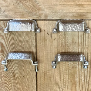May include: Four silver-coloured, hammered metal drawer pulls with a rectangular shape and decorative square accents. The pulls are mounted on a light brown wooden surface with visible grain.